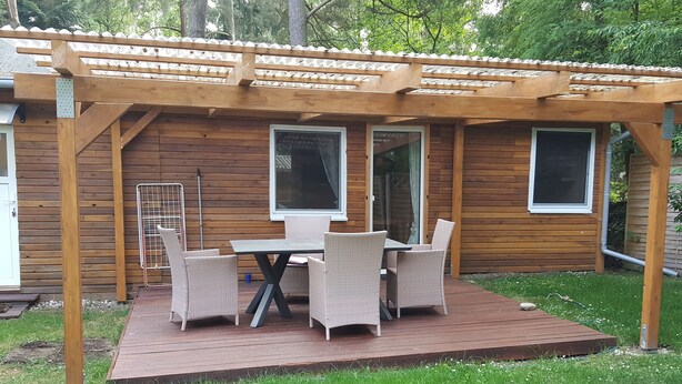 Ett trädäck med en pergola, ett bord med stolar och en gräsyta.