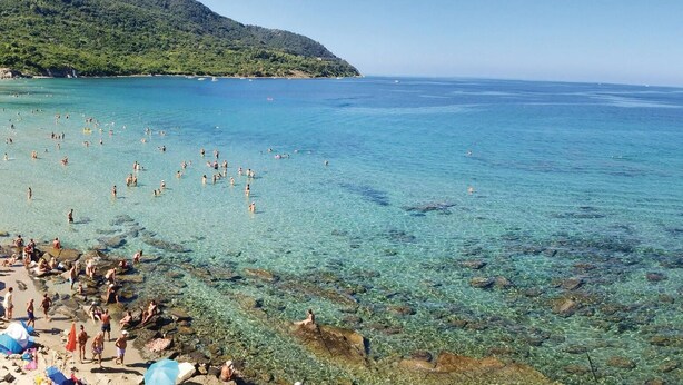 Une plage avec une eau claire et peu profonde, des zones rocheuses et un paysage montagneux en arrière-plan.