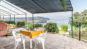 Une terrasse extérieure avec une table dressée pour deux personnes, entourée de chaises blanches, située dans un espace ombragé d’où l’on peut admirer la mer et les collines en arrière-plan.