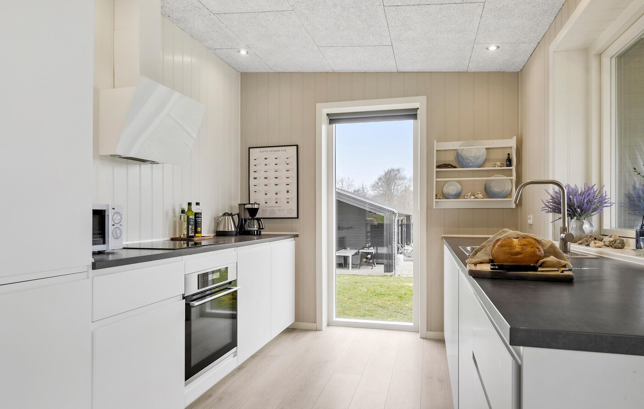 A modern kitchen with white cabinetry, a built-in oven, a coffee machine, and a large window overlooking a garden.