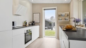Ett modernt kök med vita skåp, en inbyggd ugn, en kaffemaskin och ett stort fönster med utsikt över en trädgård.
