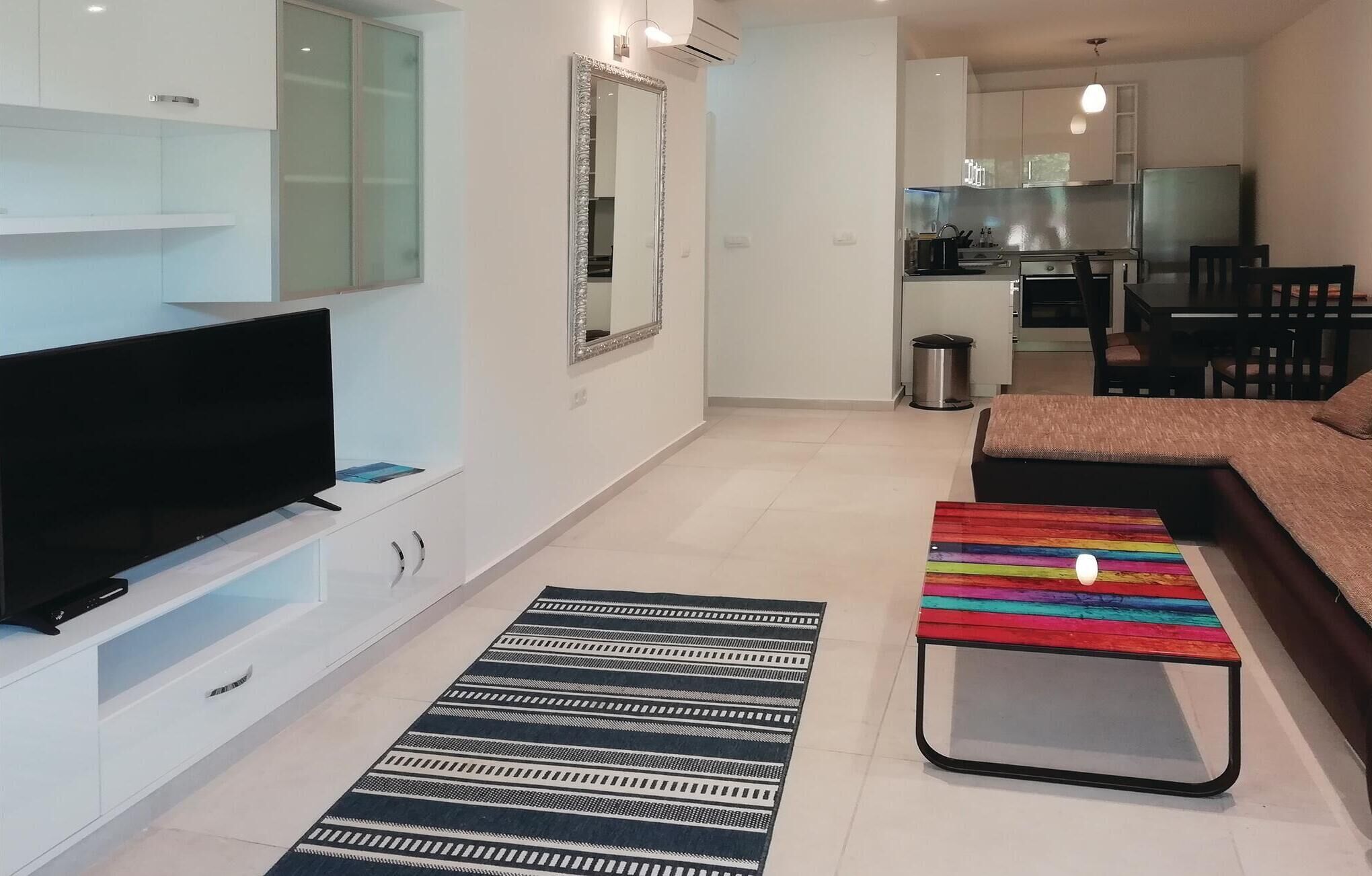 Ein modernes Wohnzimmer mit Flachbildfernseher, einem gemusterten Teppich, einem farbenfrohen Hocker und einer Couch.