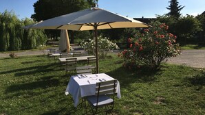 An outdoor dining area with tables and chairs under a large umbrella, surrounded by greenery and flowers.