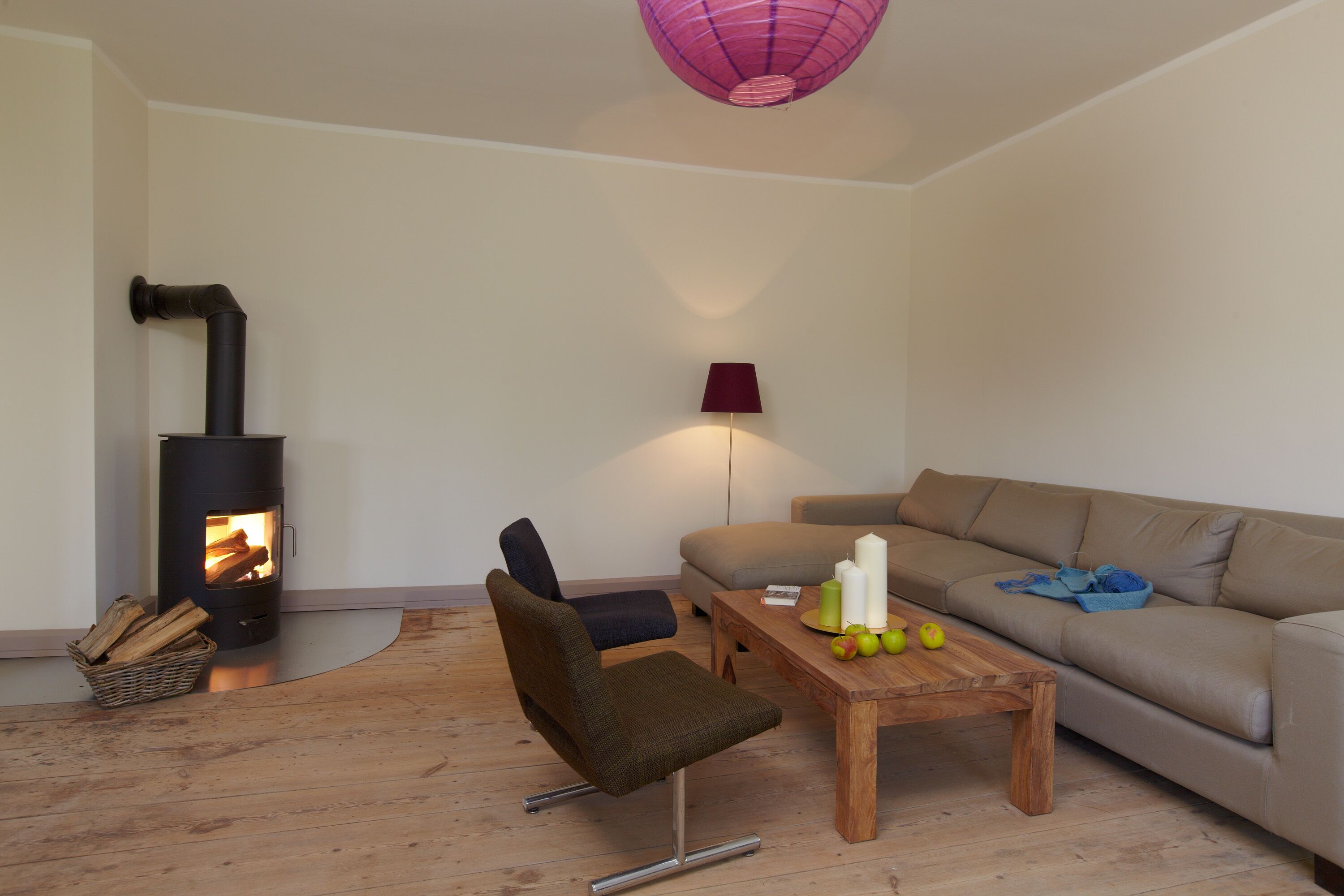 A living room with a wood-burning stove, a sofa, a chair, a coffee table with apples and candles, and a lamp.
