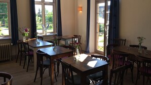 A dining area with multiple wooden tables and chairs, large windows with curtains, and a hanging light fixture.