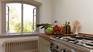 A kitchen with a stove, a window with a view of trees, a radiator, and a basket of vegetables on the counter.