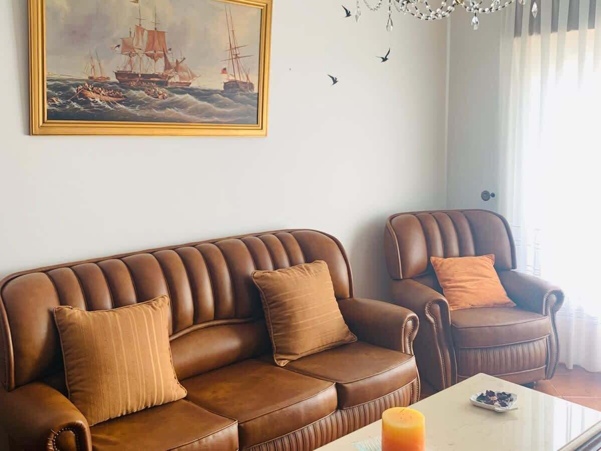 En stue med en brun lædersofa, to ens matchende lænestole, et sofabord og et maleri af et skib på væggen.