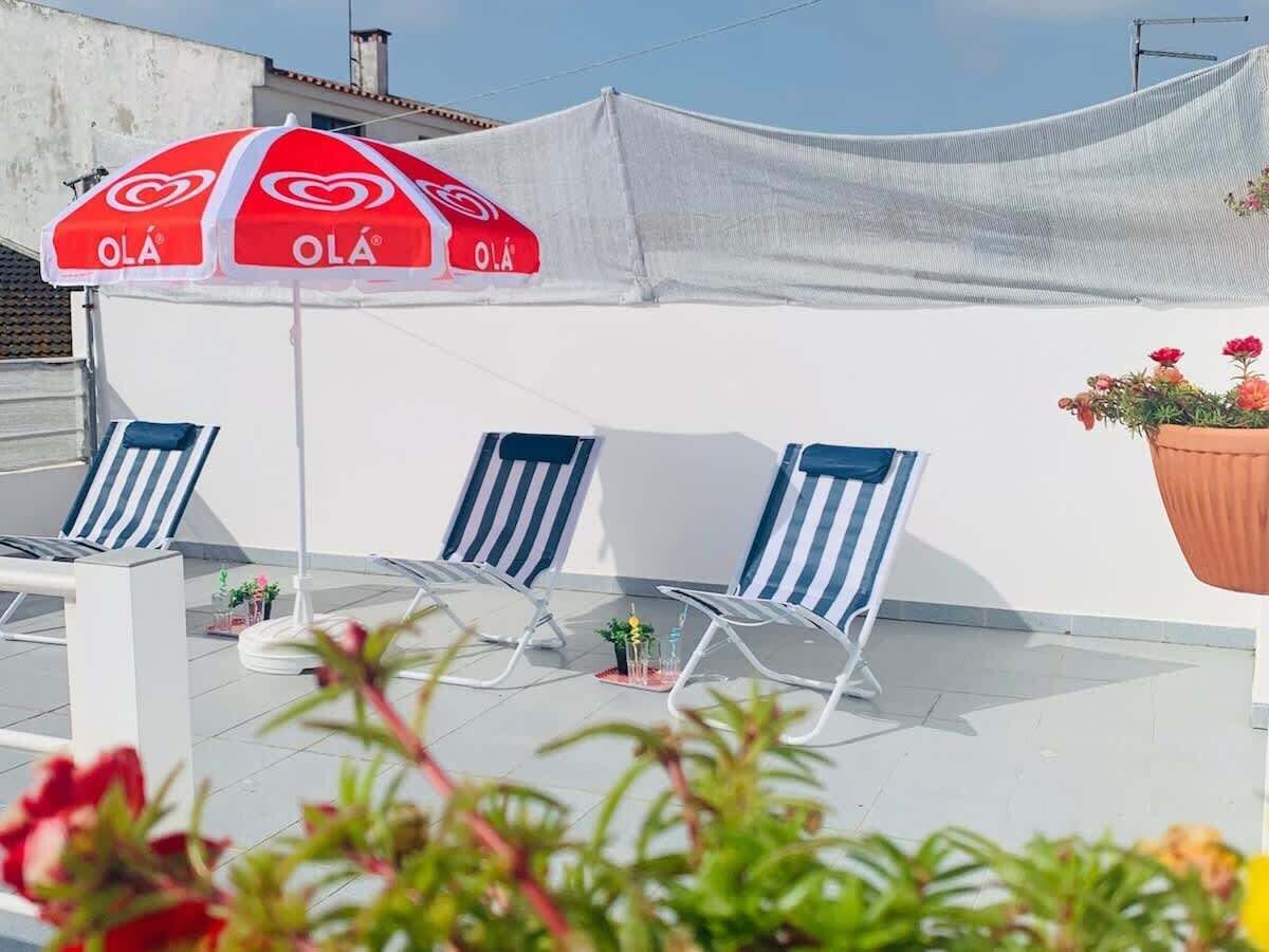 Een terras op het dak met drie gestreepte loungestoelen, een rood-witte parasol en een bloempot.
