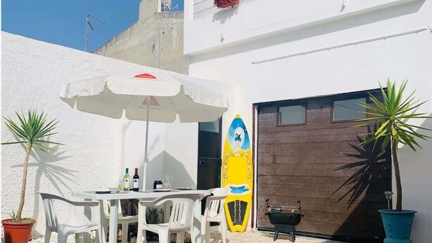 Ein weißes Gebäude mit einer Holztür, einem Tisch mit Stühlen, einem Surfbrett und Blumentöpfen.