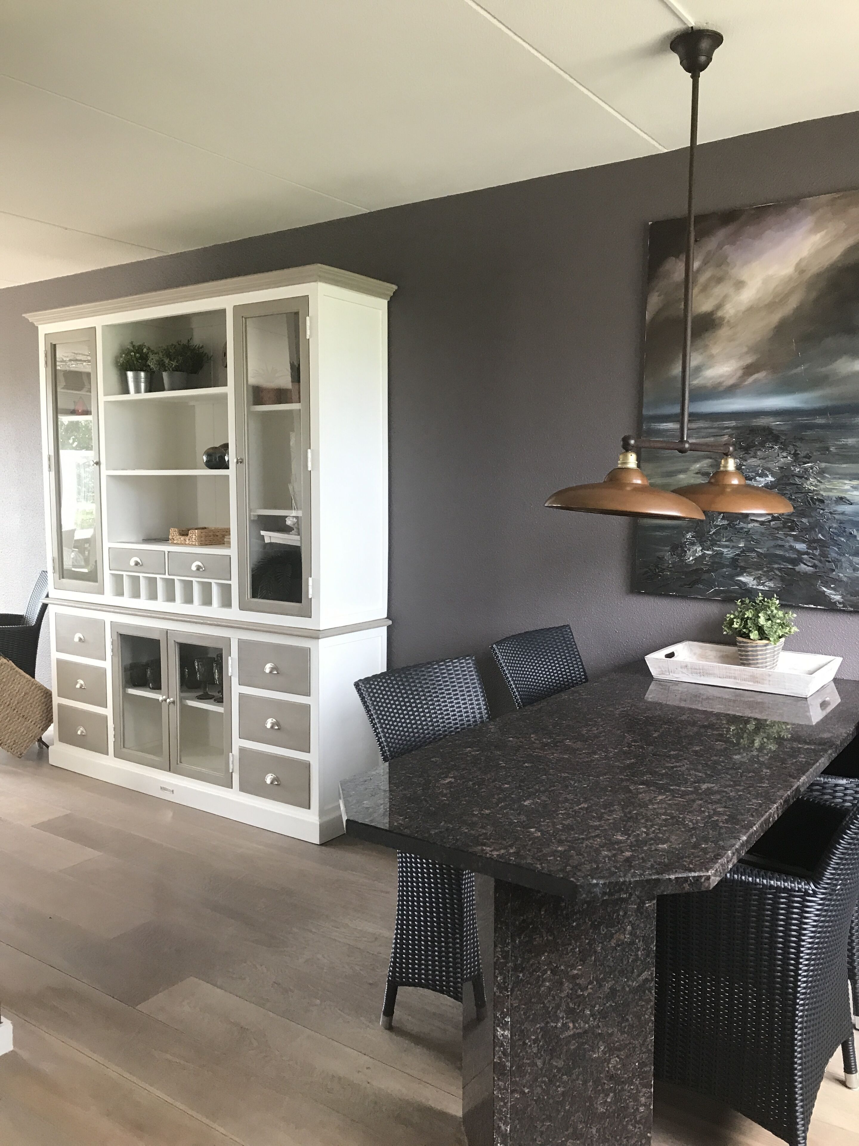 En matplats med ett mörkt bord i granit, stolar i rotting, ett vitt skåp med glasdörrar och en tavla som föreställer ett stormigt hav på väggen.