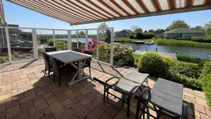 A covered patio with a table and chairs, a view of a canal, and a house in the background.