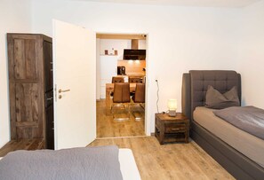 Een slaapkamer met een bed, een nachtkastje met een lamp, en een houten deur die leidt naar een keuken.
