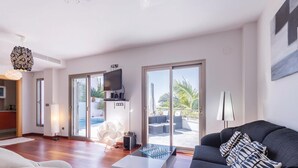Un salon moderne avec un canapé, une table basse et un téléviseur fixé au mur. Une porte-fenêtre coulissante donne accès à un espace extérieur doté d’une piscine et de mobilier de jardin.