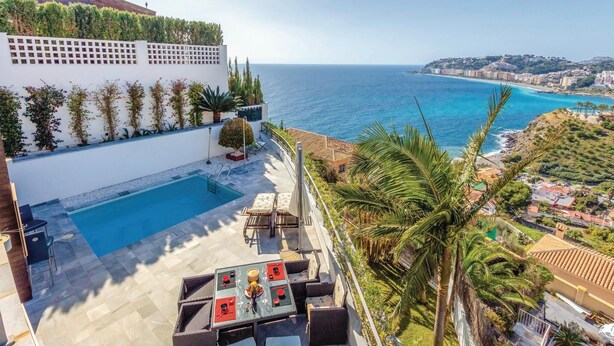 Une piscine sur le toit offrant une vue sur la mer, entourée de verdure et bordée d’un bâtiment blanc à la façade ornée d’un motif en treillis.