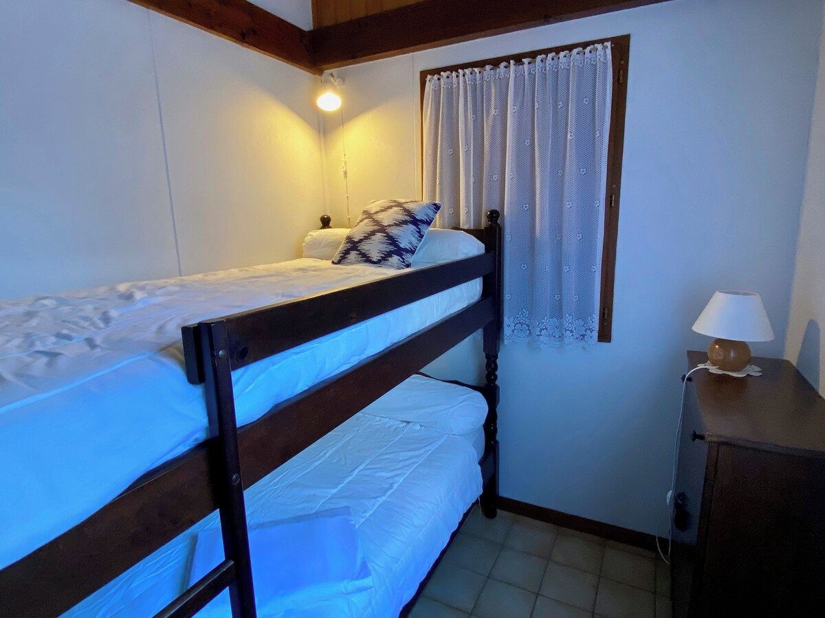 Una cama individual con cabecera, una mesita de noche con lámpara, una ventana con cortinas y una luz empotrada en la pared.