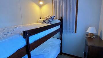 Una cama individual con cabecera, una mesita de noche con lámpara, una ventana con cortinas y una luz empotrada en la pared.