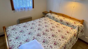 Une chambre à coucher avec un lit en bois, une literie à motifs floraux, une fenêtre ornée de rideaux en dentelle, une lampe fixée au mur et un petit tableau.