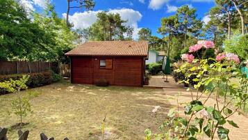 A wooden cabin with a tiled roof, surrounded by a fenced yard and a variety of plants.