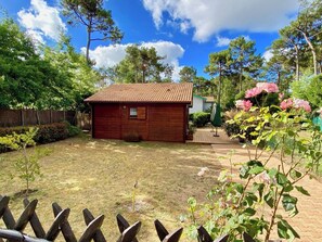 A wooden cabin with a tiled roof, surrounded by a fenced yard and a variety of plants.