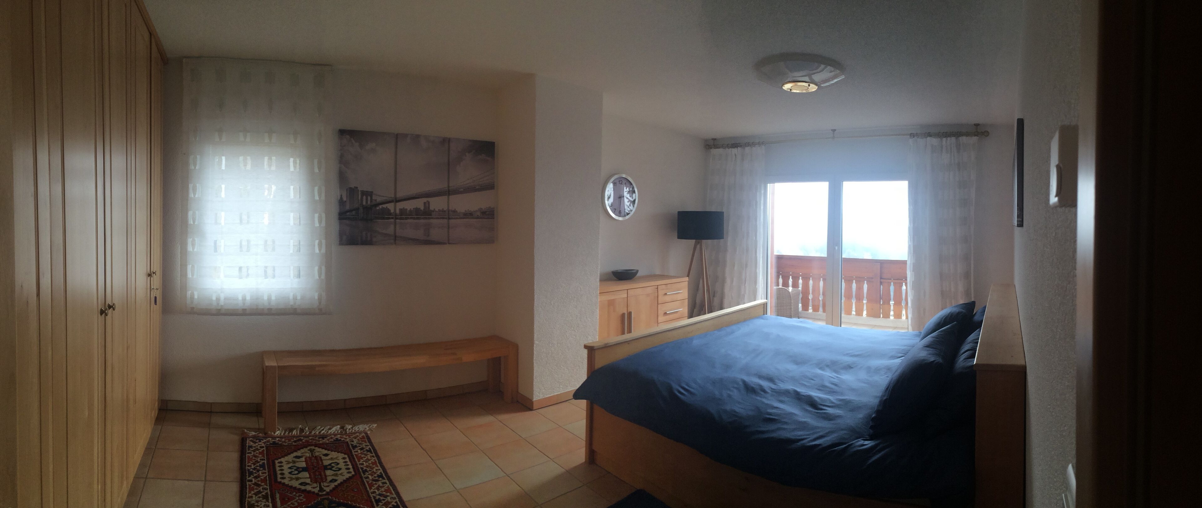 Ett sovrum med en säng, en garderob i trä, en bänk, ett litet bord med en lampa, en tavla på väggen och en balkong med utsikt.
