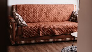 A brown quilted sofa with a plaid throw blanket, a small round table, and a patterned rug in a room with wooden flooring and a sloped ceiling.
