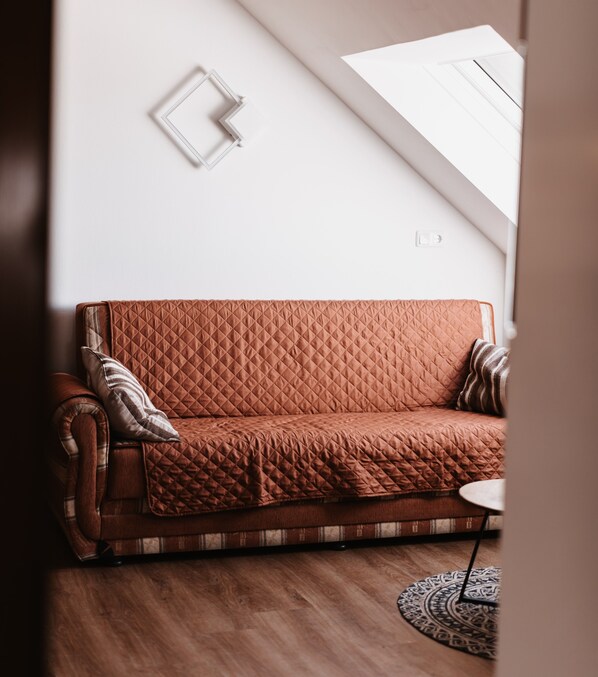 A brown quilted sofa with a plaid throw blanket, a small round table, and a patterned rug in a room with wooden flooring and a sloped ceiling.