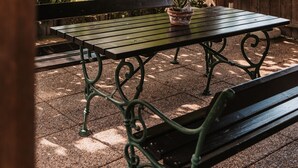 A wooden pergola with a table and benches, surrounded by greenery and a view of trees and hills.