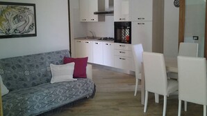 Eine moderne Küche mit weißen Schränken, einem Gasherd und einem eingebauten Backofen. Ein Essbereich mit weißen Stühlen und einem Tisch ist ebenfalls vorhanden. Im Wohnbereich steht ein graues Sofa mit gemusterten Kissen.