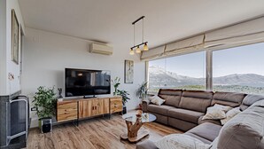 Una sala de estar moderna con un televisor de pantalla plana grande, un mueble de madera, un sofá de cuero y vistas a montañas a través de ventanas amplias.