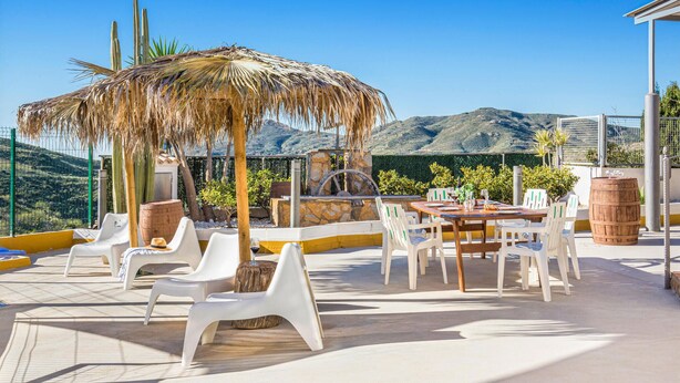 An outdoor patio with white plastic chairs and a wooden table, a thatched umbrella, and a view of mountains in the background. - Lovely home in Tallante (Tallante)
