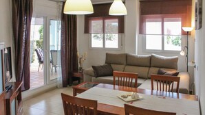 Una sala de estar con sofá, mesa de comedor y sillas. Hay ventanas con cortinas, una lámpara y un televisor.