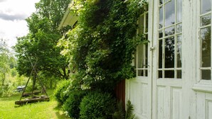 En vit byggnad med ett fönster, omgiven av frodig grönska och en trägunga.