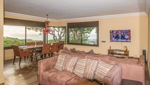 Ett vardagsrum med ett stort fönster, en matplats med flätade stolar, en rosa soffa och en tv på väggen.