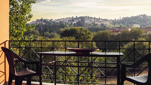 Un balcone con tavolo e sedie, affacciato su una città con edifici e alberi.