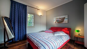 Camera da letto con un letto rosso, biancheria a righe, un quadro di una barca, una lampada da terra e una finestra con tende.