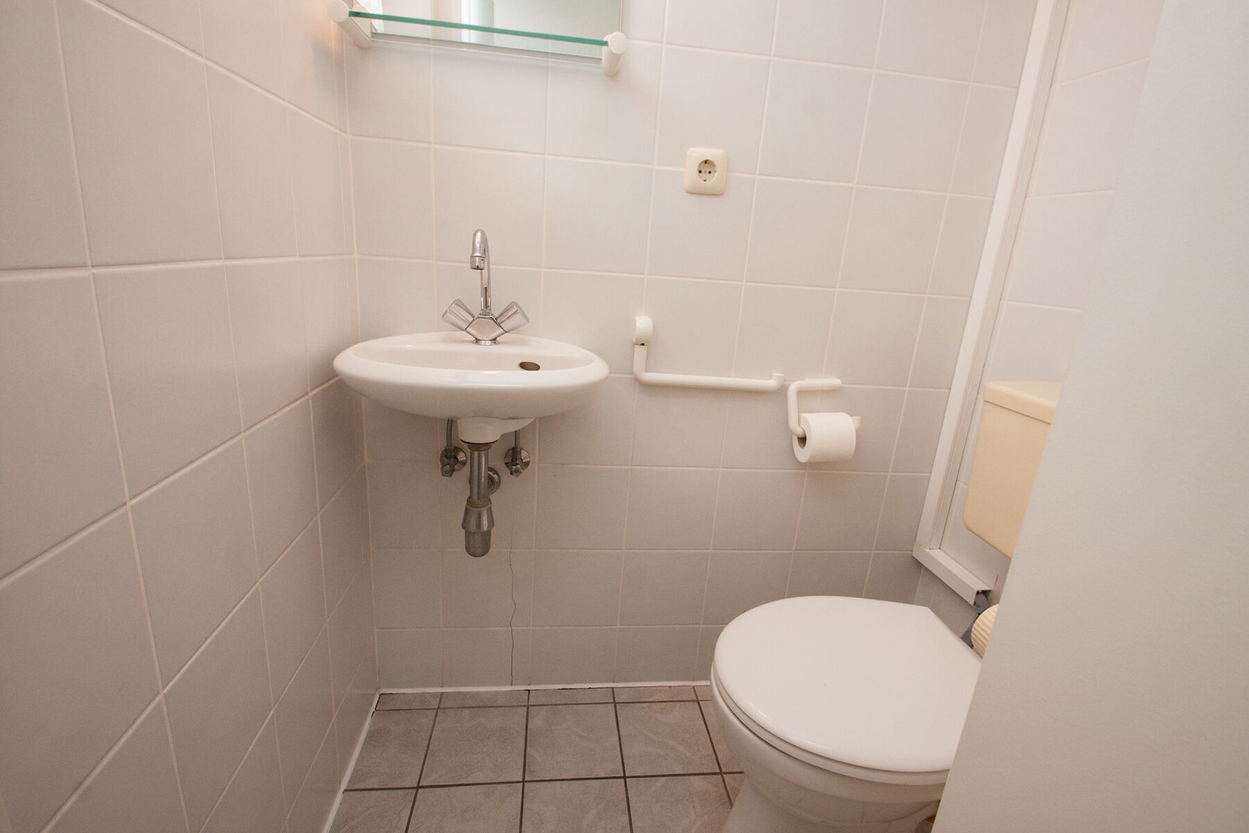 A small, clean bathroom with a white sink, toilet, and a glass shelf above the sink.