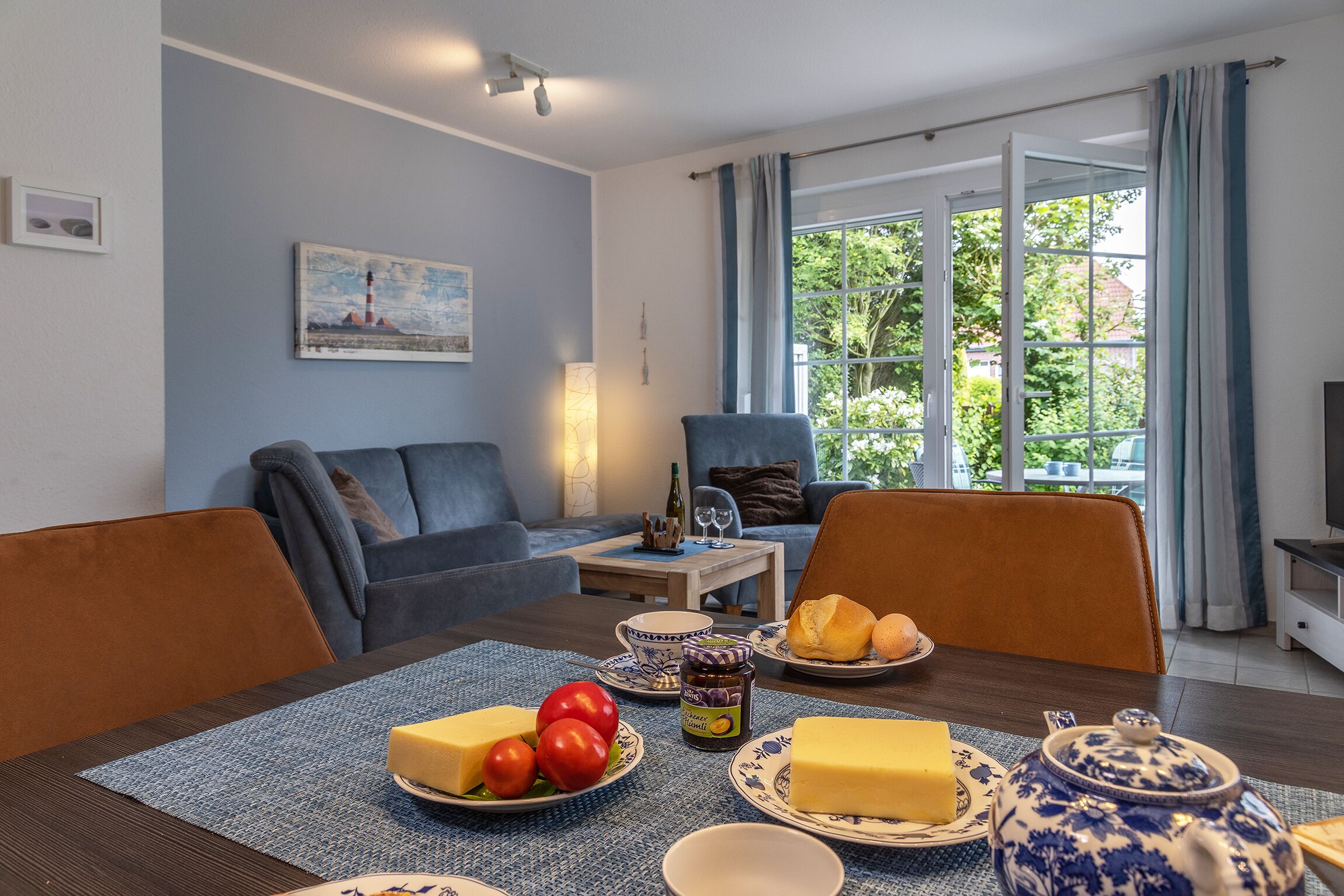 A dining area with a table set for breakfast, including cheese, tomatoes, and bread, with a view of greenery outside.
