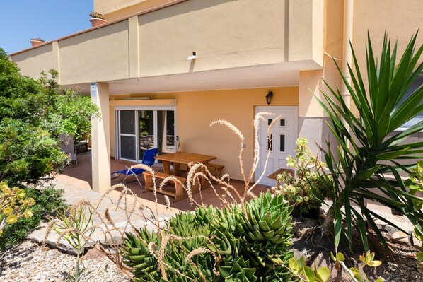A building with a patio area featuring a wooden table and chairs, surrounded by various plants and cacti.
