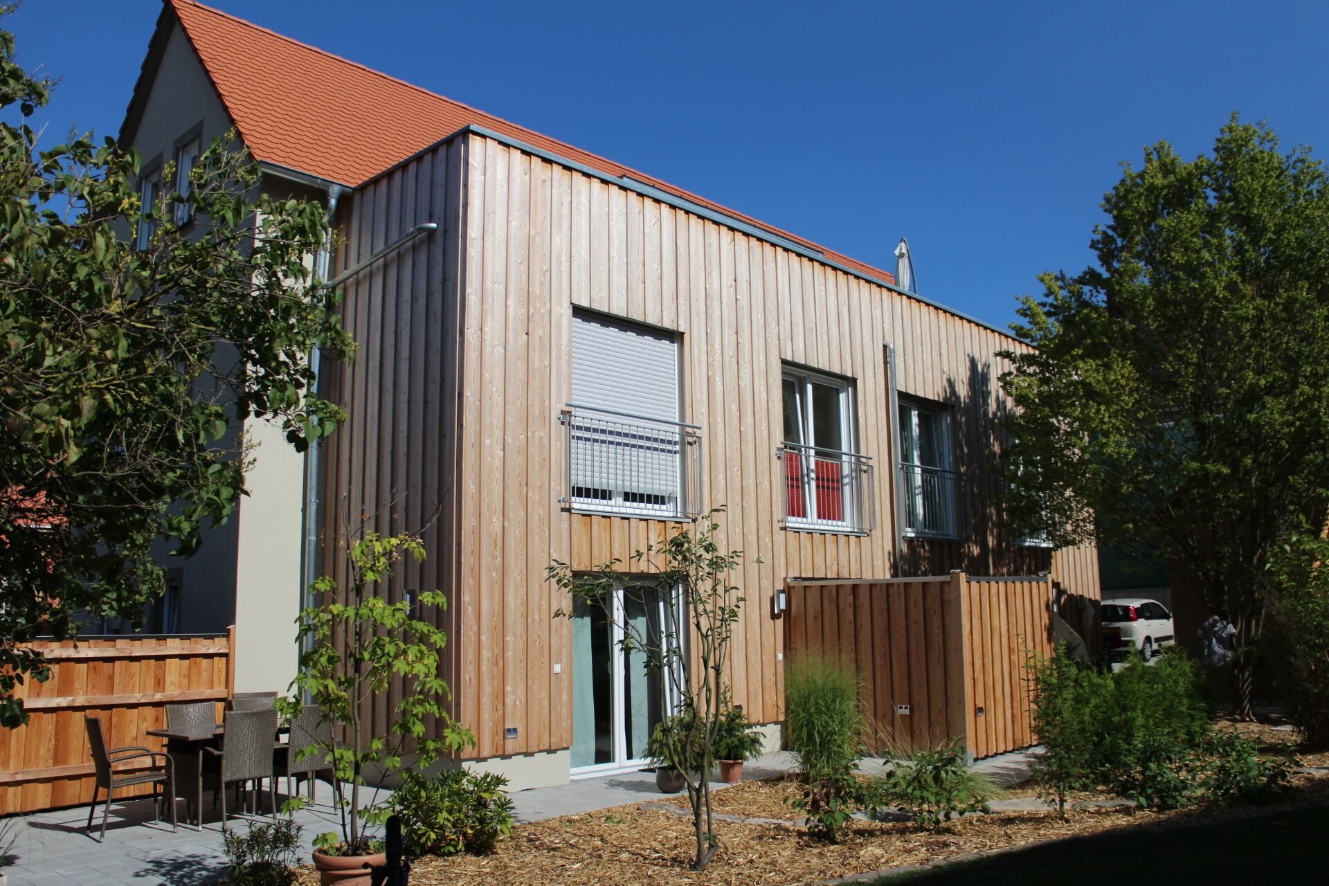 A modern two-story house with a wooden facade, a red roof, and a fenced backyard with potted plants.