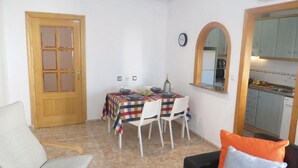 Un comedor con una mesa dispuesta para cuatro, una puerta de madera, un reloj y una cocina visible a través de un arco.
