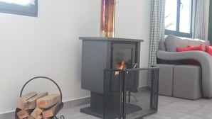 Een moderne woonkamer met een houtkachel, een stapelhout en een grijze bank met rode kussens.
