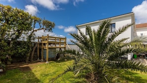 Een modern huis met een witte gevel, een houten terras en een verzorgde tuin met palmbomen.