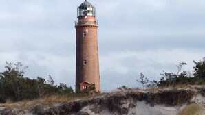 A tall brick lighthouse with a small building at its base, surrounded by sandy dunes and sparse vegetation. - small cozy apartment in a very quiet location (Prerow)