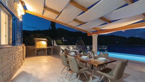 An outdoor dining area with a stone fireplace, wooden beams, and a view of a lake at dusk.