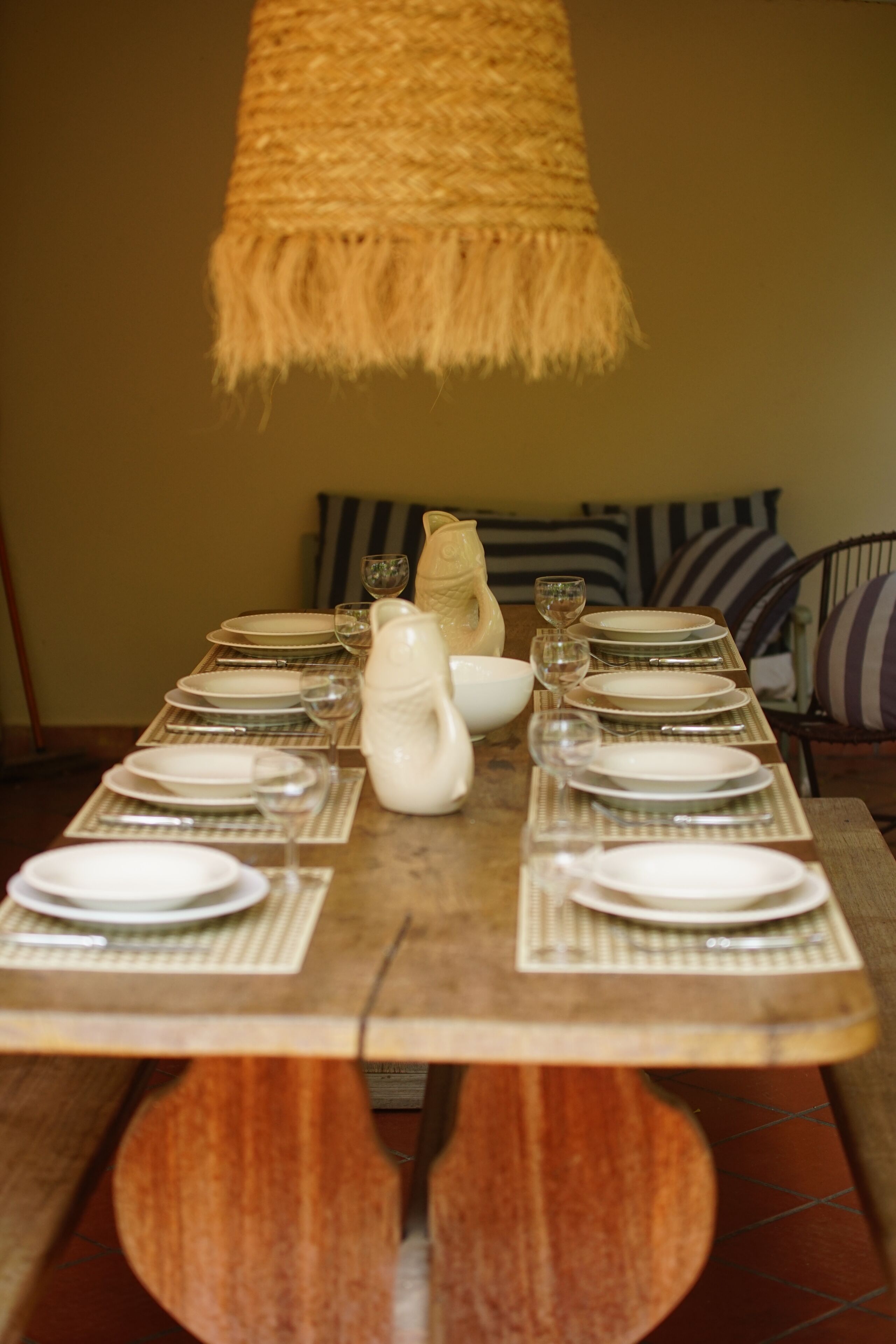 Une table à manger dressée avec des assiettes, des verres et un pichet, et une suspension au-dessus.
