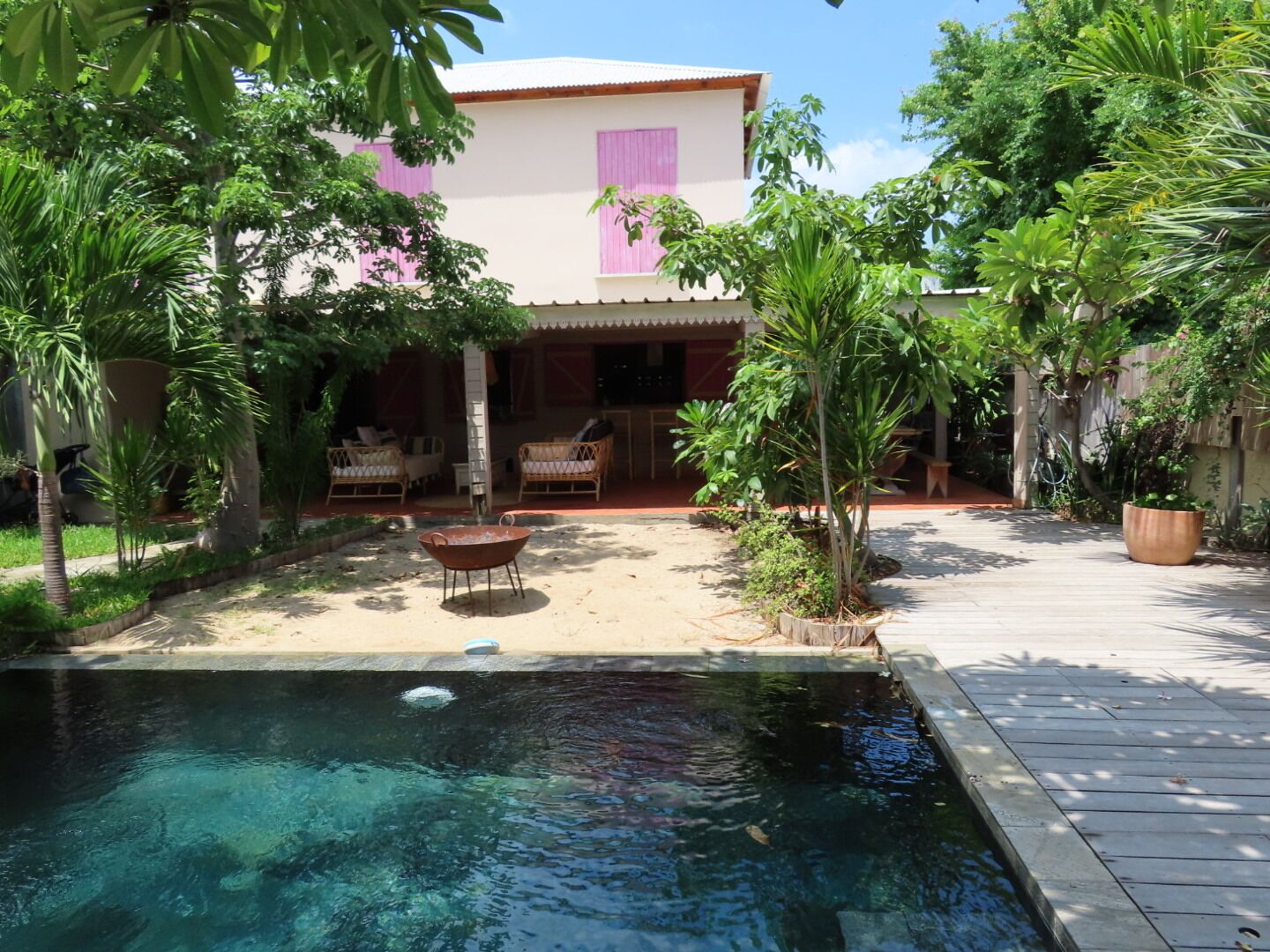 A pool with a wooden deck, surrounded by tropical plants and a building with pink shutters.