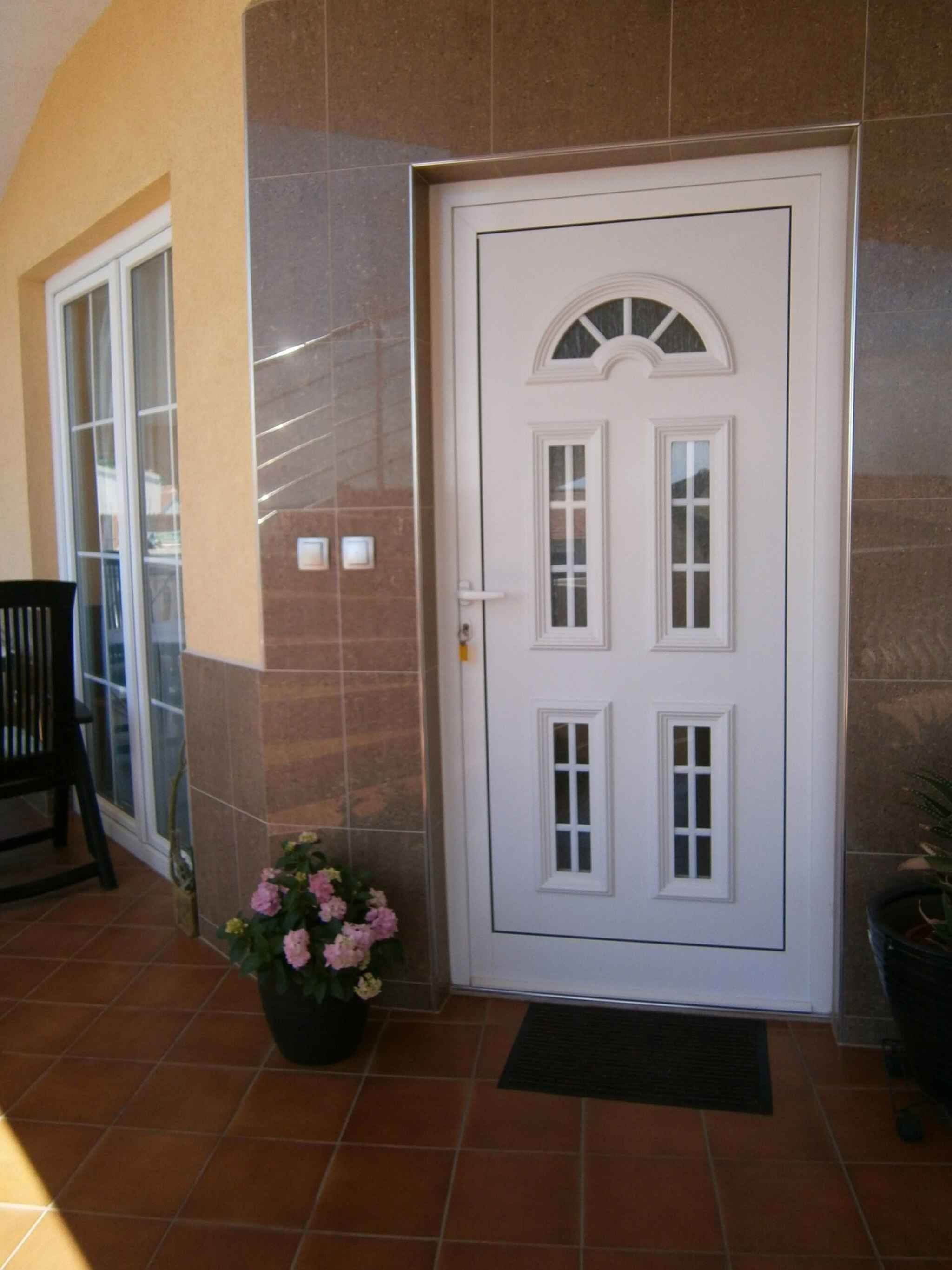 A white door with a small window, a potted plant with pink flowers, and a black mat at the entrance.
