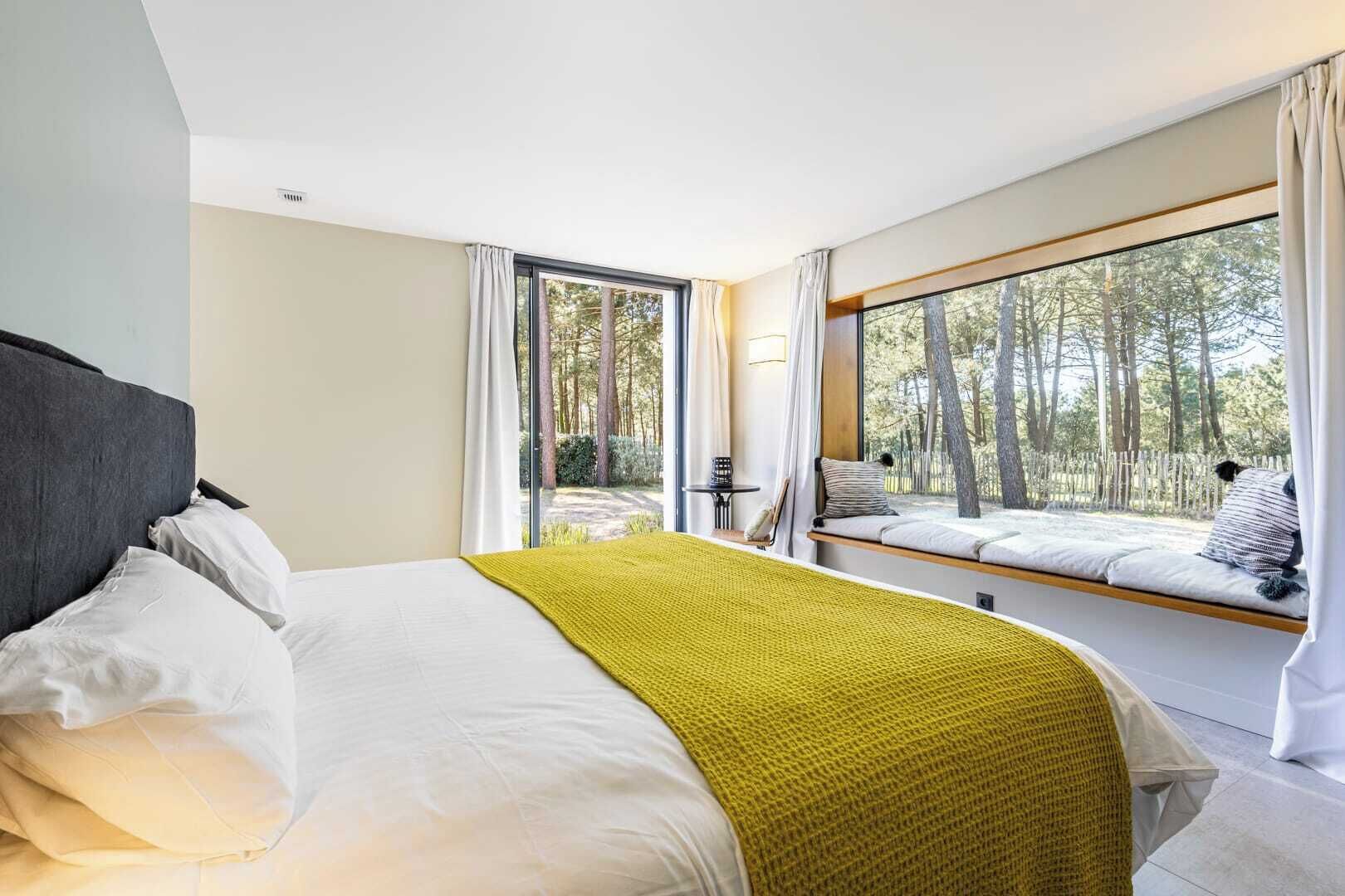 A modern bedroom with a large bed, a window seat overlooking a forest, and a view of trees.