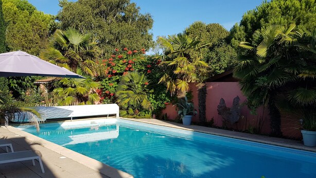 A swimming pool with a white lounge chair and umbrella, surrounded by lush greenery and a pink wall. - Guest house near downtown La Rochelle with heated pool (puilboreau)