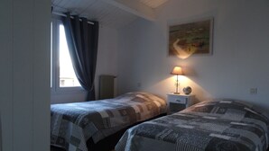 A room with two beds, a nightstand with a lamp, a painting on the wall, and a window with curtains. - Guest house near downtown La Rochelle with heated pool (puilboreau)